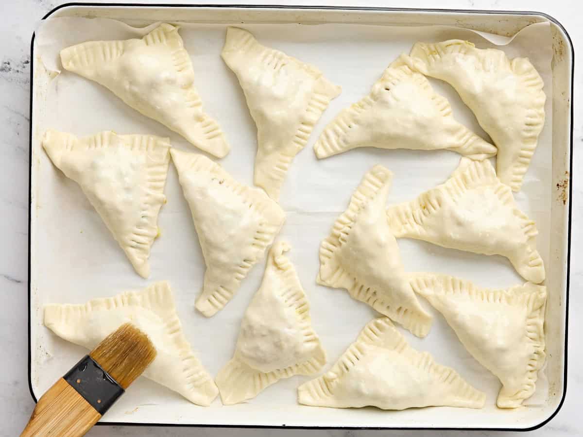 Formed homemade samosas being brushed with vegetable oil on a baking sheet.