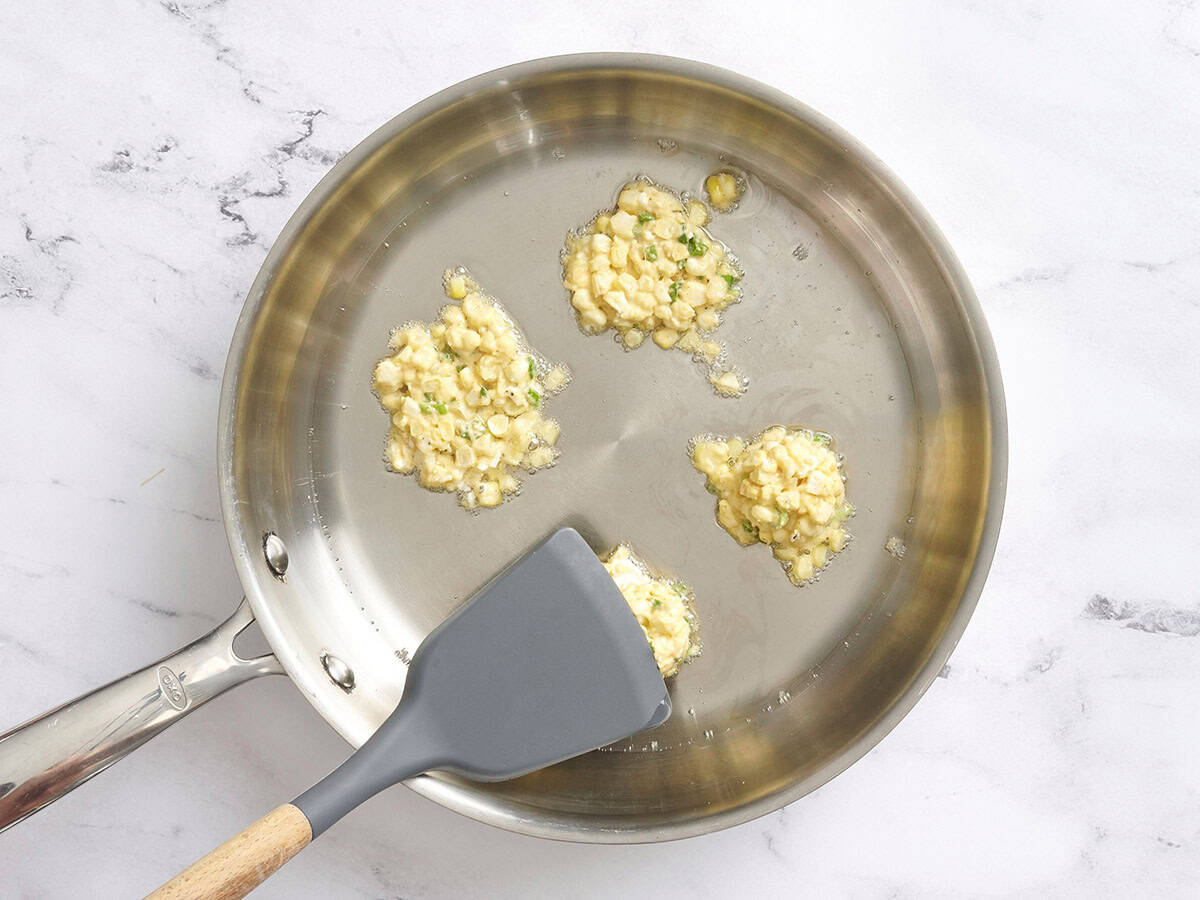 A spatula pressing down sweet corn fritters in a frying pan with oil.