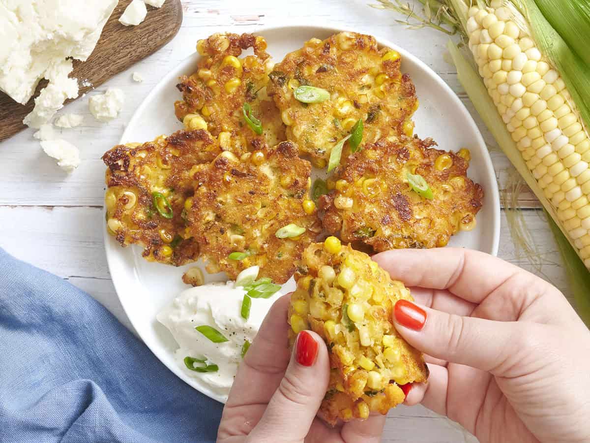 Overhead view of sweet corn fritters on a plate with sour cream and green onions, with hands breaking one fritter in half.