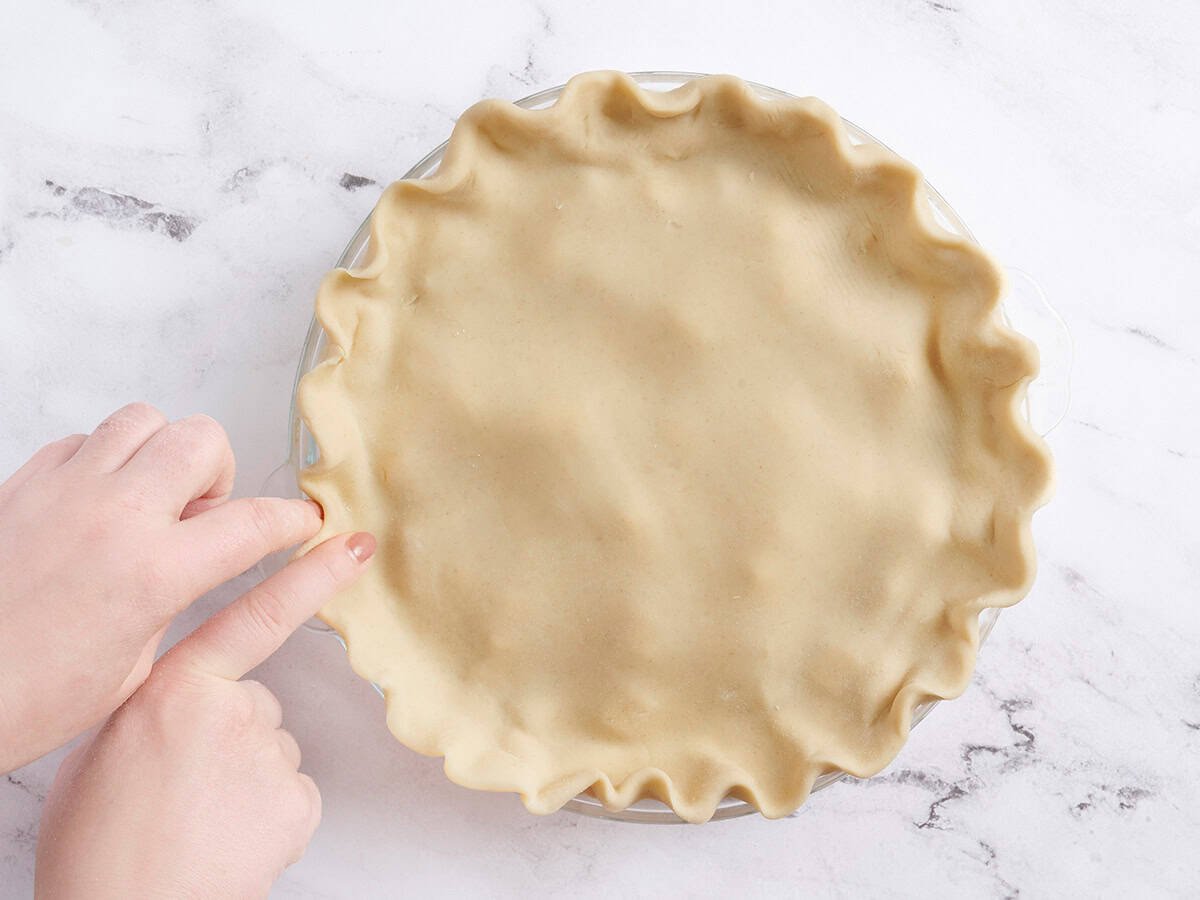 A hand crimping the top crust on a peach pie.