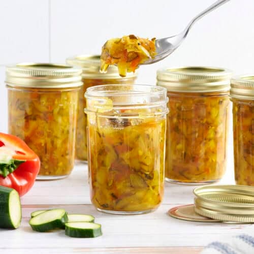 Side view of jars of zucchini relish, is a spoon taking some from one jar.