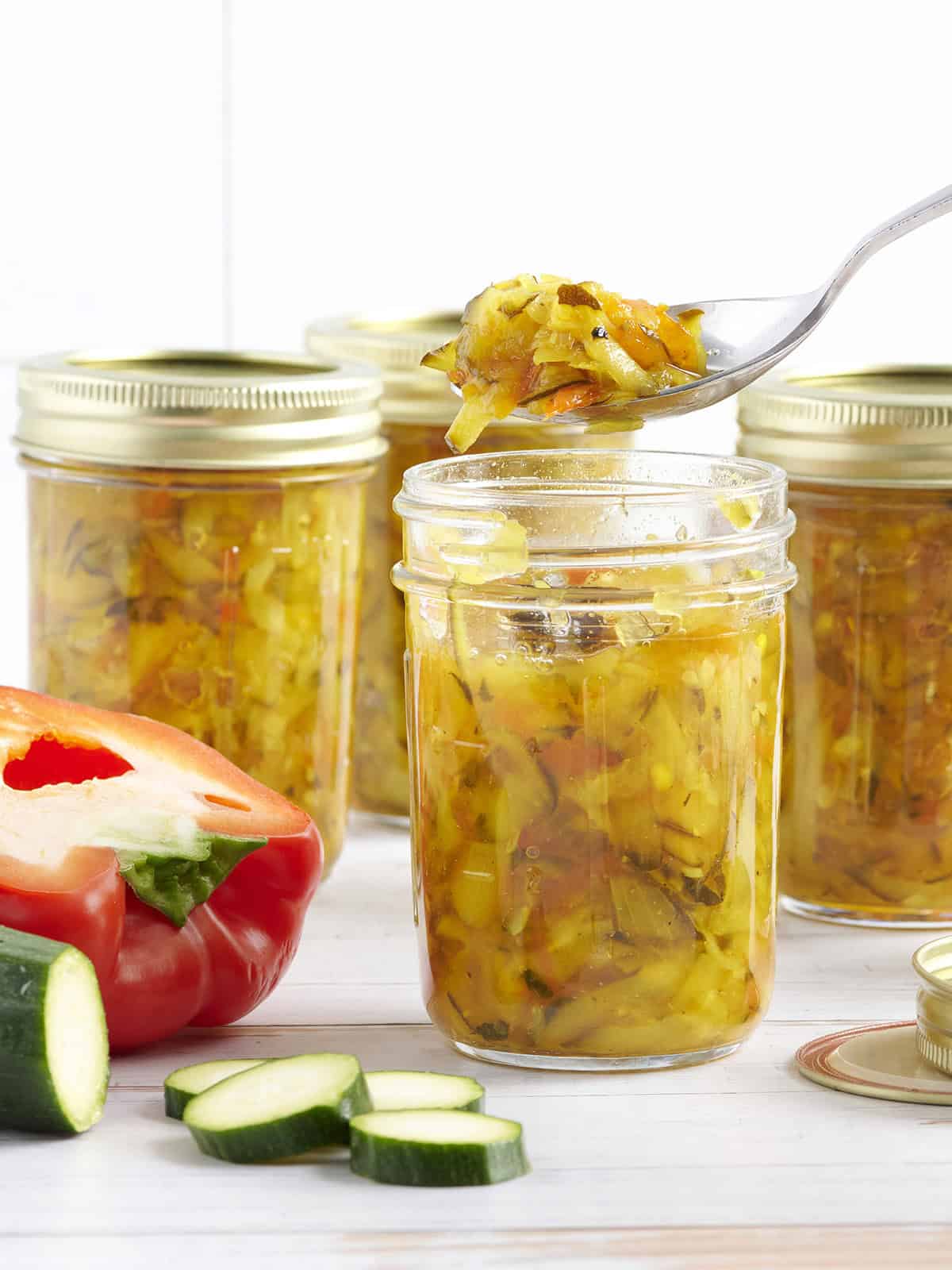 Side view of jars of zucchini relish, is a spoon taking some from one jar.