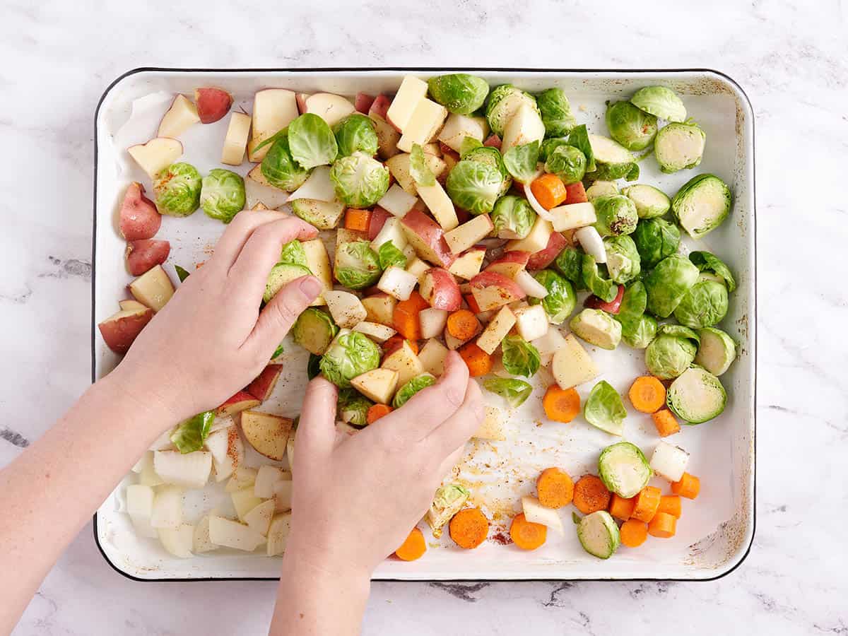Brussels sprouts, apples, red potatoes, and carrots being tossed with oil and seasonings.