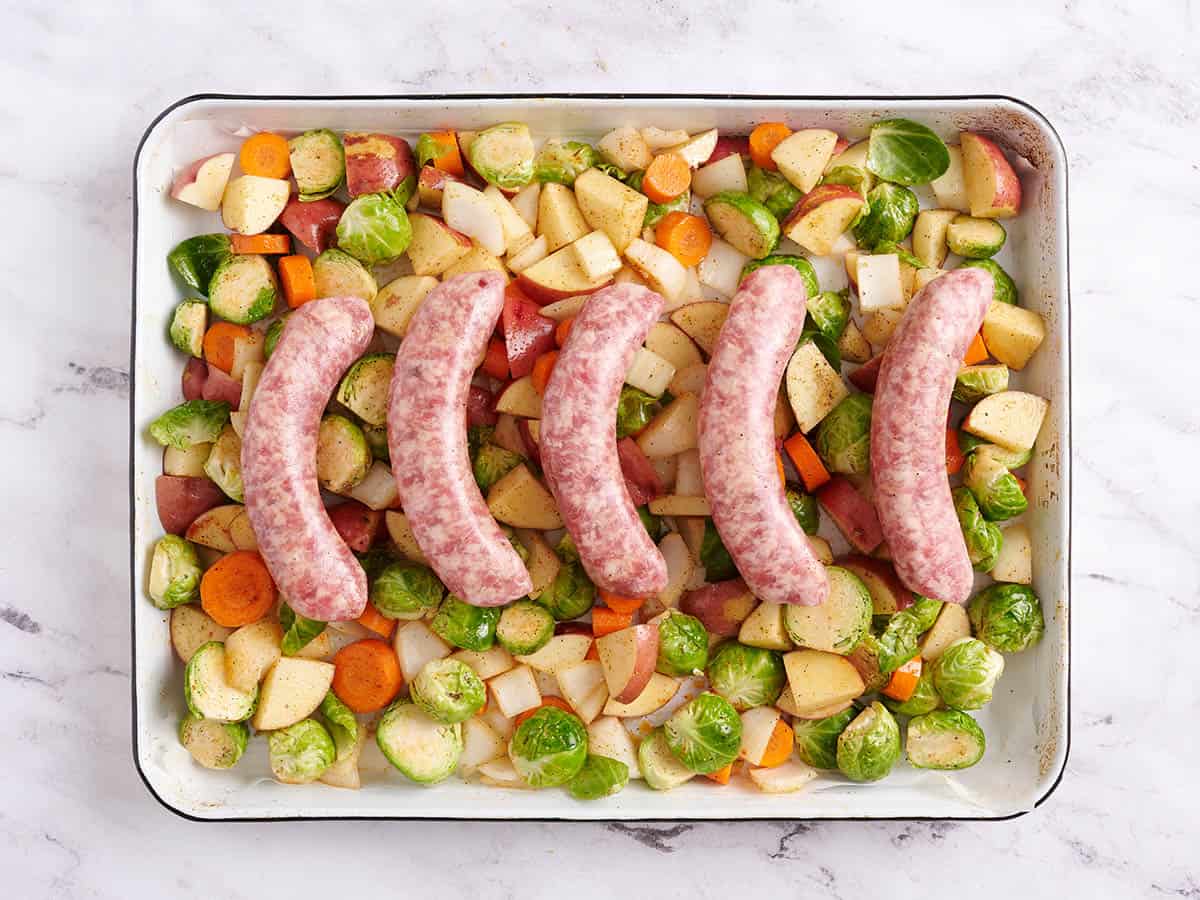 Five bratwurst on a layer of vegetables on a sheet pan.