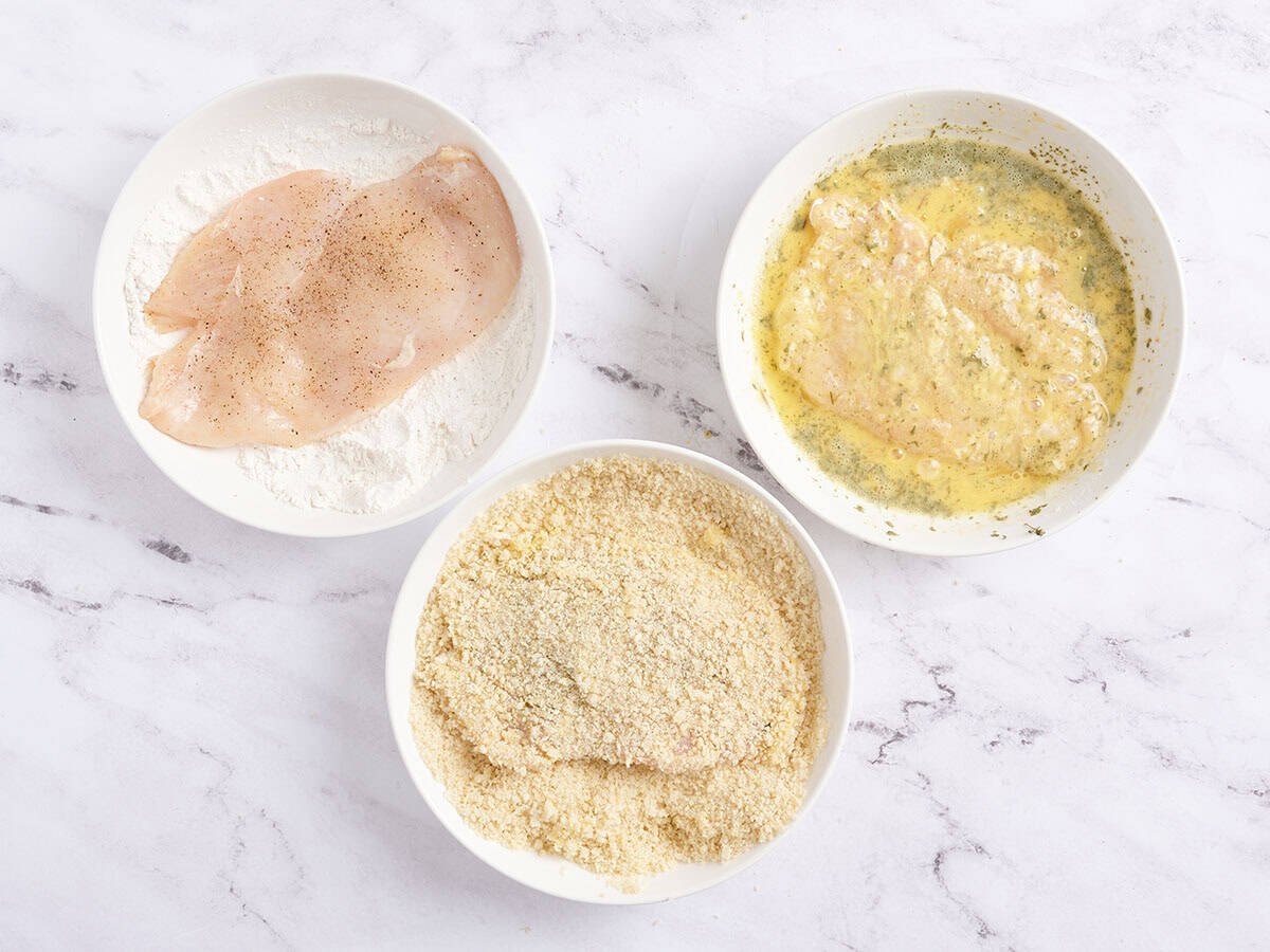 Chicken breast cutlets being dipped in flour, then egg, and then breadcrumbs in three separate bowls.