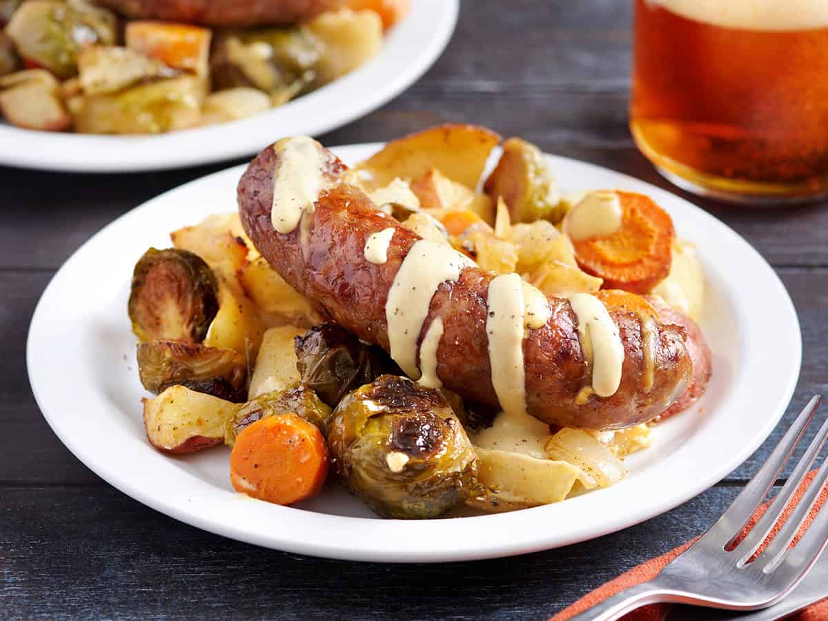 Sheet pan roasted vegetables on a plate with a bratwurst sausage, drizzled with mustard sauce.