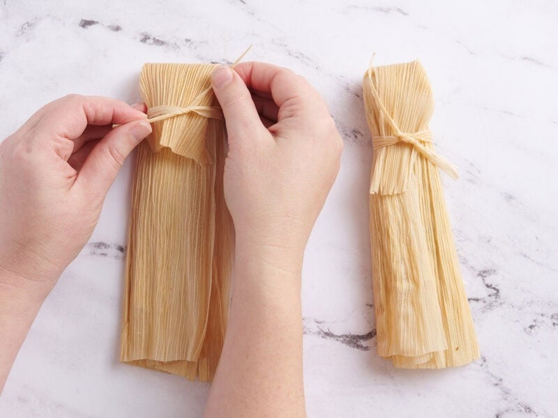 Hands tying a knot around a filled corn husk.