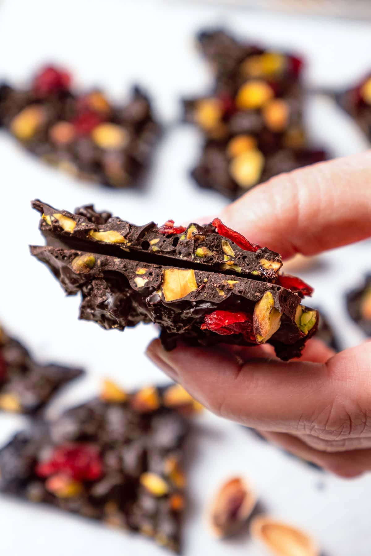 Hand holding two pieces of dark chocolate bark with pistachios and cranberries.