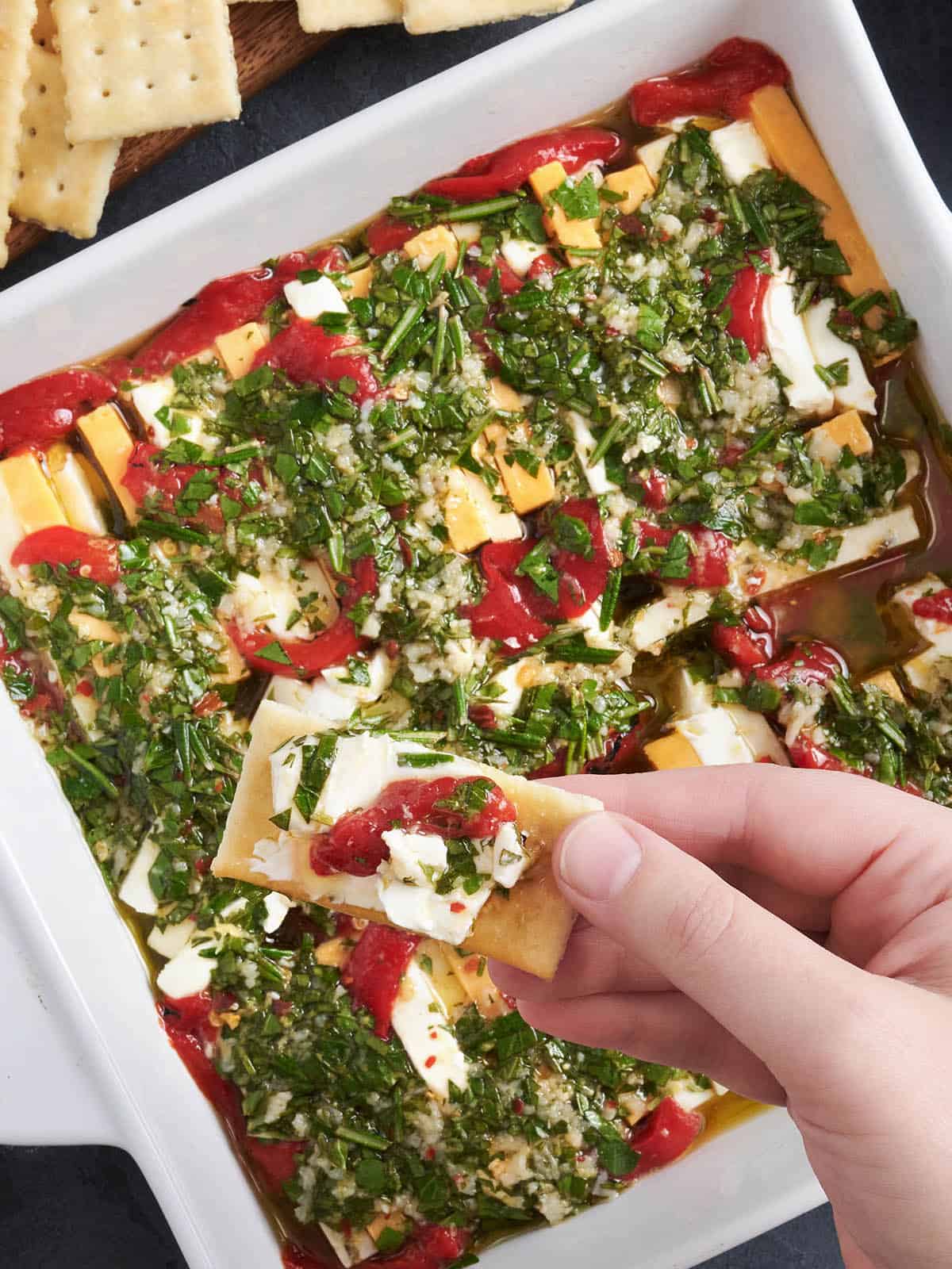 Overhead view of a pan of marinated cheese, with a hand holding a cracker with cheese on.