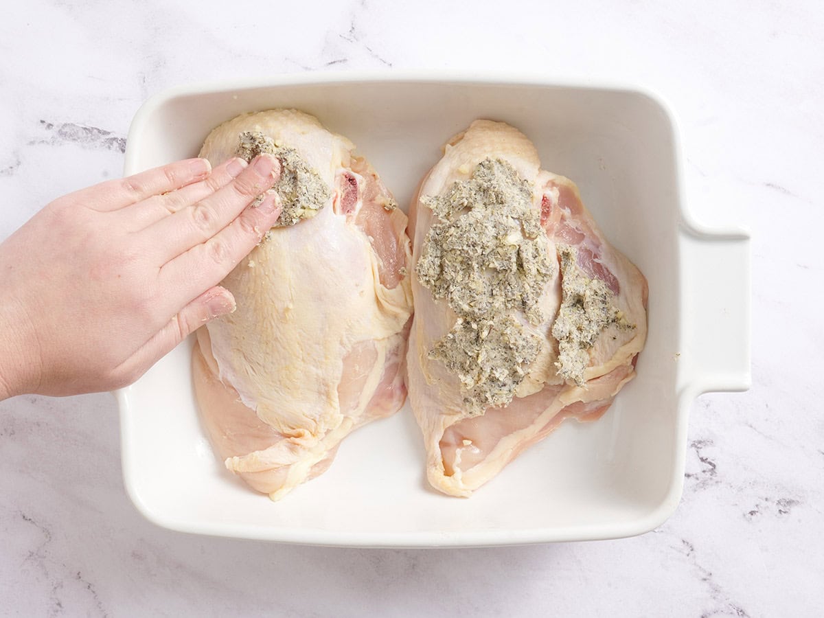 Hands rubbing butter on chicken breasts in a baking dish.